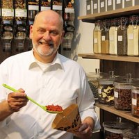 Ein Mann steht in einem Laden vor Regalen voller Gewürze, Nüsse und getrockneter Lebensmittel und füllt rote Trockenbeeren mit einem grünen Messlöffel in eine braune Papiertüte.