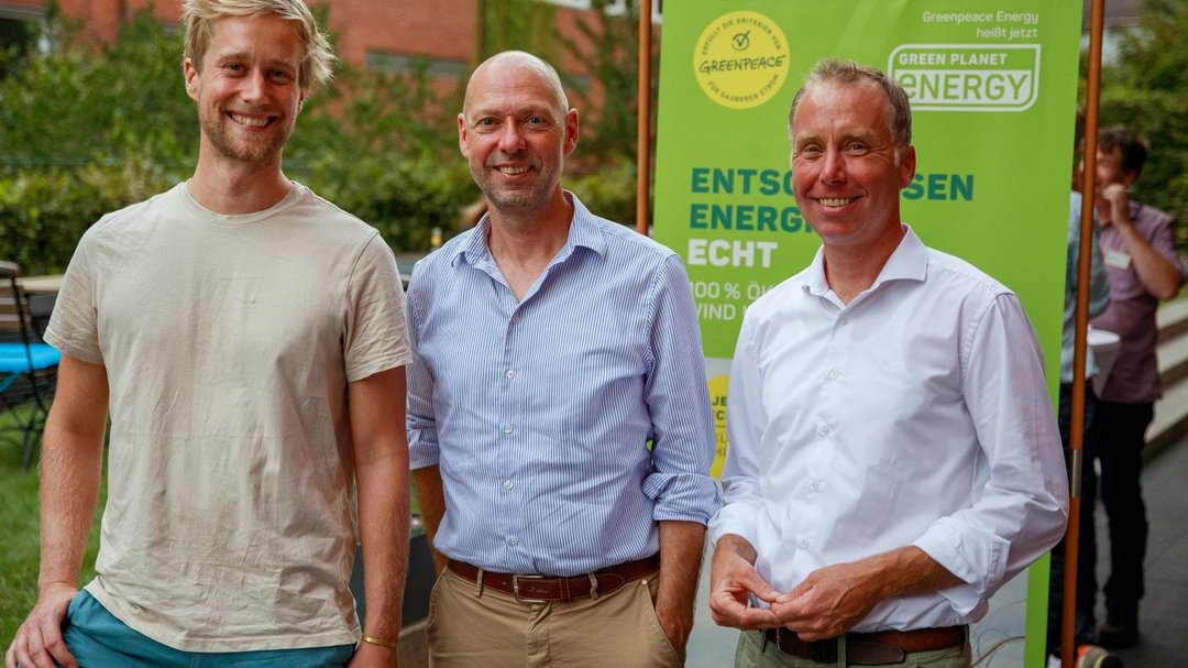 Drei Männer stehen lächelnd nebeneinander im Freien vor einem grünen Banner mit der Aufschrift „Greenpeace Energy heißt jetzt Green Planet Energy. Entschlossen. Energie. Echt.“