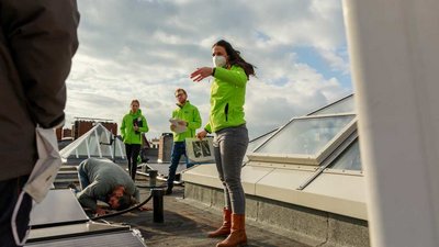 Eine Gruppe von Menschen in leuchtend grünen Jacken inspiziert und installiert Solarzellen auf einem Dach. Eine Frau gibt Anweisungen, während ein Mann sich über die Paneele beugt. Der Himmel ist teils bewölkt.