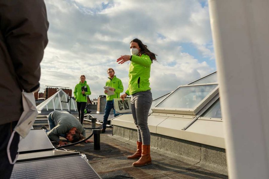 Eine Gruppe von Menschen in leuchtend grünen Jacken inspiziert und installiert Solarzellen auf einem Dach. Eine Frau gibt Anweisungen, während ein Mann sich über die Paneele beugt. Der Himmel ist teils bewölkt.