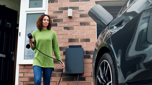 Eine Person hält ein Ladekabel und geht in Richtung eines Elektroautos, das vor einem Haus mit Backsteinfassade steht. Im Hintergrund ist eine Ladestation an der Wand angebracht.