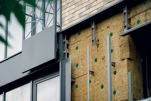 Wand mit Isolierung und Fassadenelementen, Detailansicht eines Gebäudes in Bauphase, zeigt Dämmmaterial und Metallrahmen, umgeben von Ziegelstruktur.