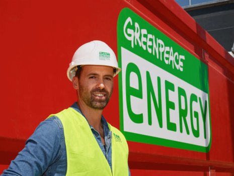 Ein Arbeiter mit Bauhelm und Warnweste steht vor dem Greenpeace Energy Logo.
