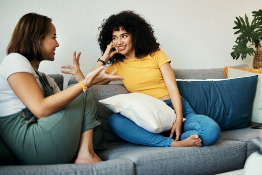 Zwei Frauen mit dunklen Haaren sitzen auf einem Sofa und unterhalten sich gestikulierend und lächelnd. Im Umfeld stehen Pflanzen und ein weißer Couchtisch.