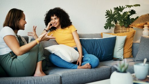 Zwei Frauen mit dunklen Haaren sitzen auf einem Sofa und unterhalten sich gestikulierend und lächelnd. Im Umfeld stehen Pflanzen und ein weißer Couchtisch.