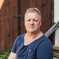 Jens Hauser (54), Landwirt und Anti-Braunkohle-Aktivist aus Pödelwitz in Sachsen Eine Person sitzt auf einer Bank, trägt ein dunkelblaues T-Shirt, und befindet sich vor einem Holztor. Streiflicht von links sorgt für einen sonnigen Tag.