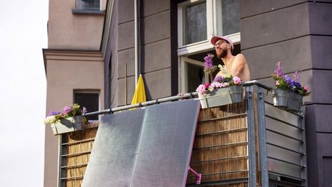 Eine Person steht ohne Oberbekleidung auf einem Balkon. Der Balkon ist mit Blumen geschmückt und an der Vorderseite ist ein großes Solarpanel angebracht.