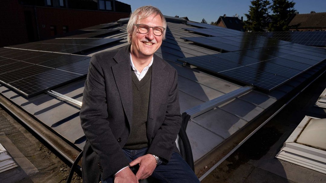 Ein lächelnder Mann mit grauen Haaren, grauem Schnurrbart und Brille sitzt auf der Kante eines Dachs auf dem eine PV-Anlage montiert ist