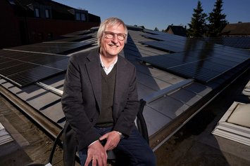Ein lächelnder Mann mit grauen Haaren, grauem Schnurrbart und Brille sitzt auf der Kante eines Dachs auf dem eine PV-Anlage montiert ist