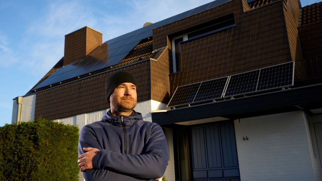 Ein Mann mit schwarzer Mütze und verschränkten Armen steht im Sonnenlicht und blickt leicht lächelnd an der Kamera vorbei. Im Hintergrund ist ein Haus mit PV-Modulen zu sehen.