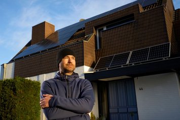 Ein Mann mit schwarzer Mütze und verschränkten Armen steht im Sonnenlicht und blickt leicht lächelnd an der Kamera vorbei. Im Hintergrund ist ein Haus mit PV-Modulen zu sehen.