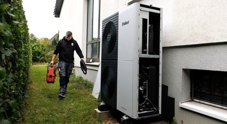 Ein Mann in Arbeitshose und schwarzem Hoodie trägt einen roten Werkzeugkoffer und einen schwarzen Schlauch in Richtung einer Vaillant-Wärmepumpe, die an der Seite eines Hauses steht.