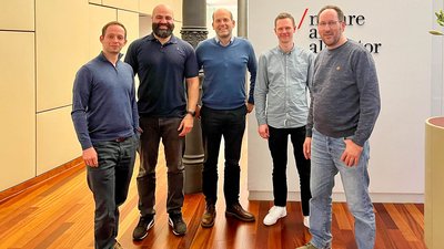 Fünf Männer stehen lächelnd in einem modernen Büro auf Holzfußboden. Im Hintergrund sind beige Wände und ein Schild mit teilweise erkennbarem Text: „nature a“ sichtbar.