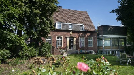 Ein zweistöckiges Backsteinhaus mit Giebeldach steht in einem Garten. Im Vordergrund blühen Rosen, umgeben von grüner Vegetation und Bäumen. Klarer, blauer Himmel im Hintergrund.