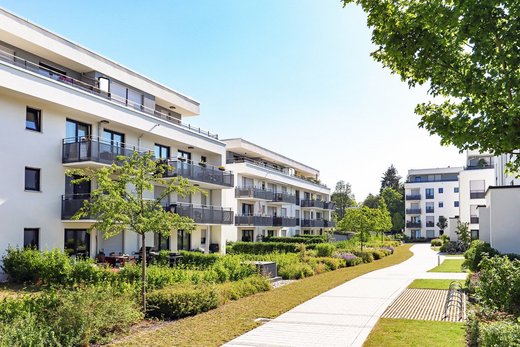 Mehrstöckige Wohngebäude stehen links und rechts eines gepflegten Weges, der durch begrünte Anlagen mit Bäumen und Büschen führt. Der Himmel ist klar und sonnig.