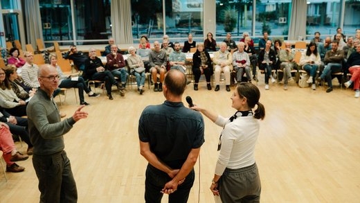 Zwei Personen von hinten mit einem Mikrofon stehen vor einer Gruppe von Menschen, die auf Stühlen in einem Halbkreis sitzen.