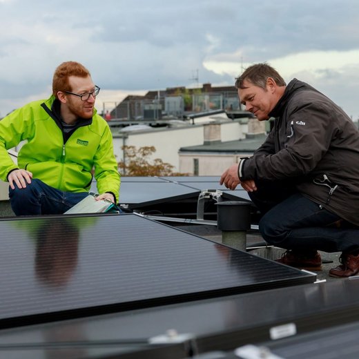 Zwei Männer knien auf einem Flachdach inmitten von Solaranlagen und begutachten diese. Einer trägt eine grüne Jacke mit Green Planet Energy Logo und hält Zettel in der Hand.