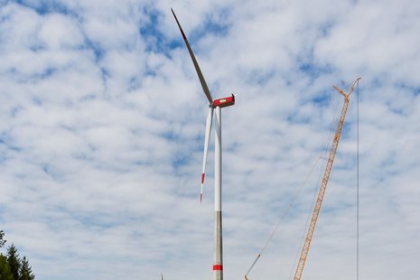 Windkraftanlage in der Mitte mit einem gelben Kran auf der rechten Seite und einem teilweise bewölkten Himmel im Hintergrund.