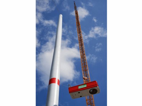 Ein riesiger Kran hebt ein Rotorblatt empor. Der Himmel ist blau mit vereinzelten Wolken. Text auf dem Kran: „VESTAS“.