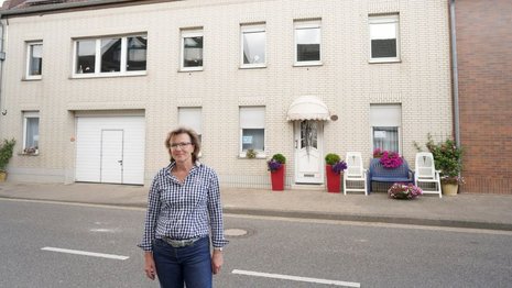 Eine Frau mit blonden, mittellangen Haaren steht auf einer Straße vor einem Haus und lächelt. Sie trägt ein blau kariertes Hemd und blaue Jeans.