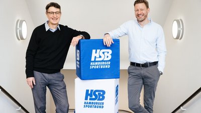 Bernard Koessler vom Hamburger Sportbund und Nils Müller von Green Planet Energy stehen im Treppenhaus neben blauen und weißen Würfeln, auf denen das Logo des Hamburger Sportbundes zu sehen ist.