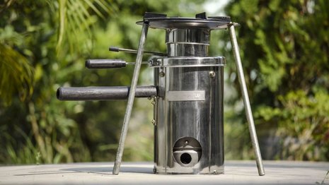 Ein Edelstahlkocher steht mit Griffen und Stützbeinen auf einem Tisch im Freien, umgeben von grüner Vegetation. Keine Textelemente sind im Bild vorhanden.