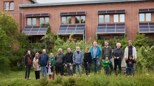 Eine Gruppe unterschiedlich alter Menschen, darunter auch Kinder, steht vor einem Mehrfamilienhaus. Über den Fenstern und am Dach sind Solarpaneele angebracht.