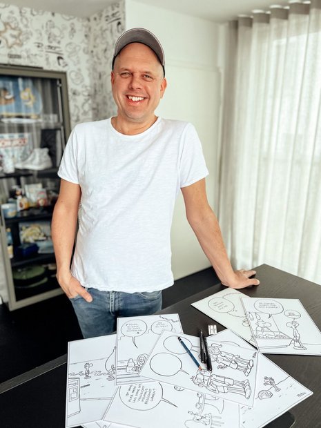Mann in weißem T-Shirt mit einer Cap steht hinter einem Schreibtisch mit Zeichnungenn und guckt in die Kamera