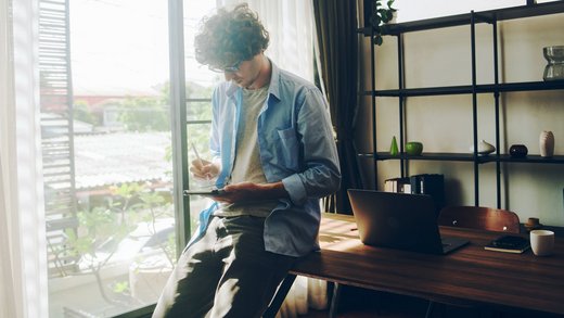 Junger Mann in grauem T-Shirt und offenem blauen Hemd lehnt an einem Schreibtisch und hält ein Tablet und einen Stift.