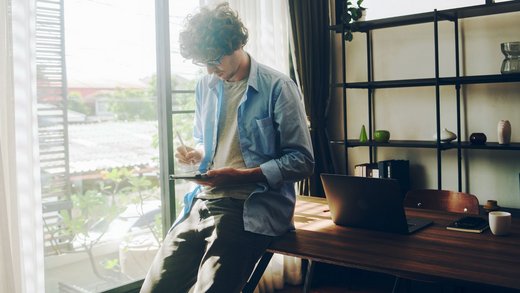 Junger Mann in grauem T-Shirt und offenem blauen Hemd lehnt an einem Schreibtisch und hält ein Tablet und einen Stift.