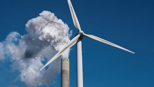 Eine Windkraftanlage und ein industrieller Schornstein mit Rauchschwaden vor einem blauen wolkenfreien Himmel.