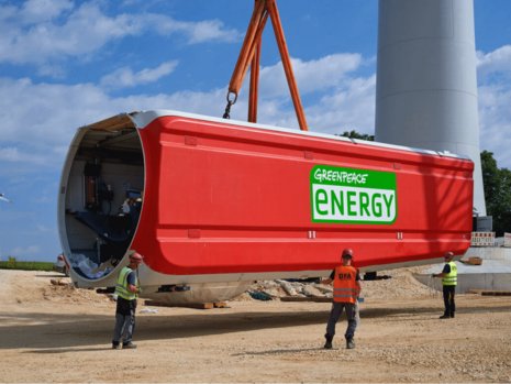 Ein großes, rotes Bauteil einer Windkraftanlage mit dem Text "GREENPEACE ENERGY" wird von einem Kran angehoben. Drei Arbeiter in Sicherheitskleidung stehen daneben auf einer Baustelle.