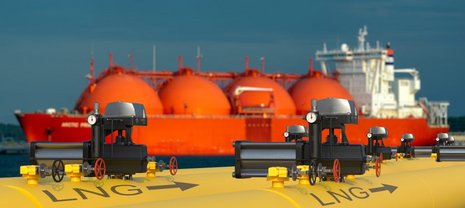Gelbe LNG-Pipelines mit Ventilen und Druckmessern im Vordergrund, während ein großes, rotes Tankschiff im Hintergrund im Hafen liegt. Das Schiff transportiert verflüssigtes Erdgas. Klarer Himmel.