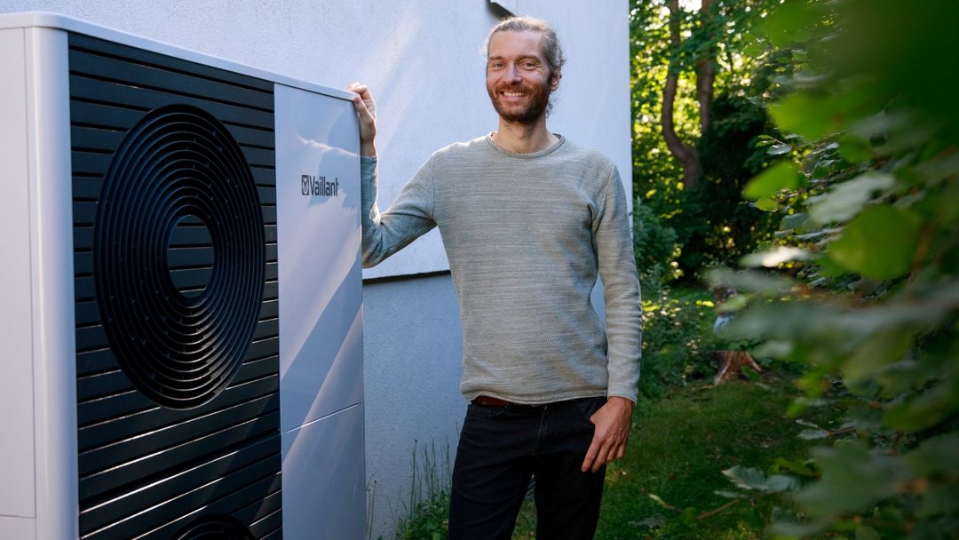 Ein Mann mit grauen Haaren und Zopf steht neben einem großen Kasten (Wärmepumpe). Im Hintergrund ist ein Haus und Garten zu sehen.
