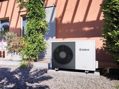 Eine installierte Wärmepumpe liefert einem mit Pflanzen umgebenen Bestandsgebäude Wärmepumpenstrom.