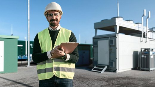 Mann mit weißem Bauhelm, gelber Warnweste und Klemmbrett in den Armen steht vor einer Art Container. Das Umfeld sieht industriell und technisch aus. Im Hintergrund sind Windräder zu sehen.