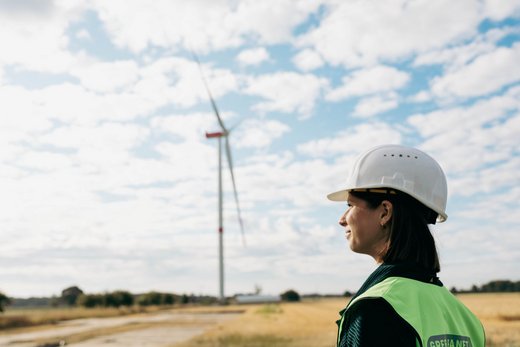 Eine Frau mit schulterlangen braunen Haaren trägt einen weißen Bauhelm und eine gelbe Warnweste. Sie steht auf einem Stoppelfeld vor einer Windkraftanlage. Der Himmel ist blau bewölkt.