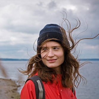 Bild einer jungen braunhaarigen Frau am Wasser. Ihre Haare wehen ihr ins Gesicht. Sie trägt eine rote Jacke, einen Rucksack und eine blaue Beanie-Mütze.