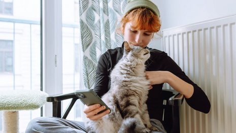 Eine Frau sitzt mit Handy in der einen Hand im Schneidersitz auf einem Stuhl vor einer Heizung und streichelt mit der anderen Hand eine Katze auf ihrem Schoß.