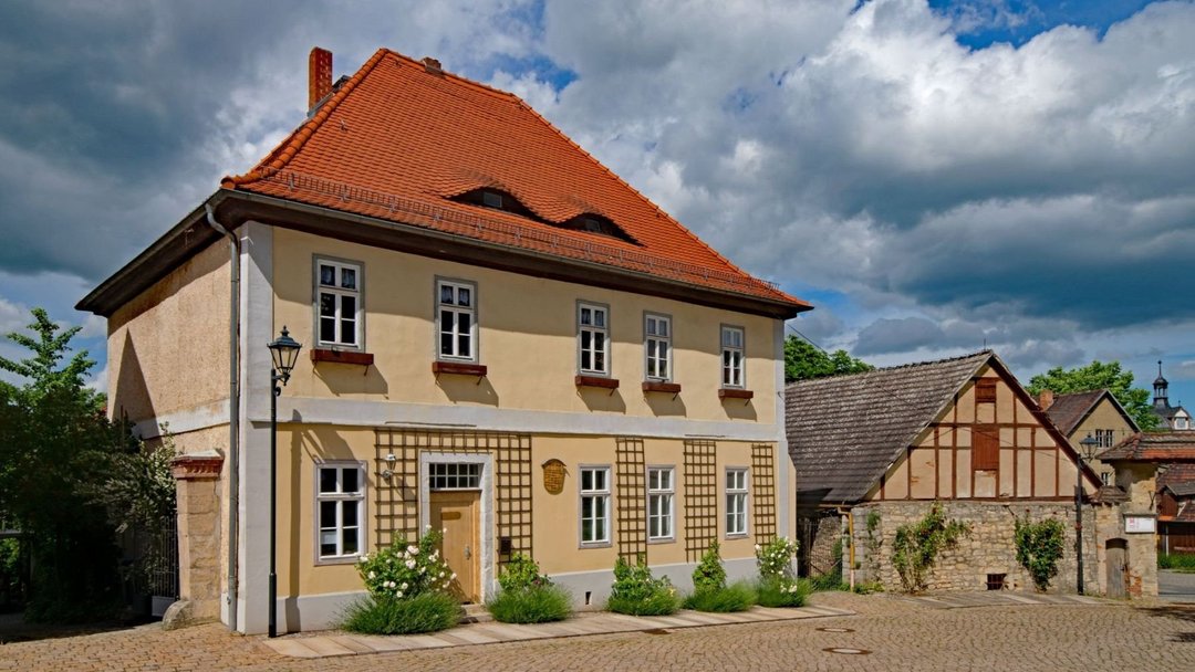Älteres sandfarbenes Haus an einer Art Marktplatz. Das Haus hat kleine Fenster mit leeren Blumenkübeln davor. An der Hauswand sind Rankhilfen angebracht. Neben der Tür ist eine hölzerne beschriftete Tafel angebracht.
