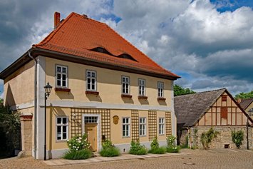 Älteres sandfarbenes Haus an einer Art Marktplatz. Das Haus hat kleine Fenster mit leeren Blumenkübeln davor. An der Hauswand sind Rankhilfen angebracht. Neben der Tür ist eine hölzerne beschriftete Tafel angebracht.