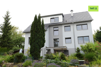 Modernes Mehrfamilienhaus mit grauer Fassade. Davor sind viele grüne Pflanzen und Büsche gepflanzt.