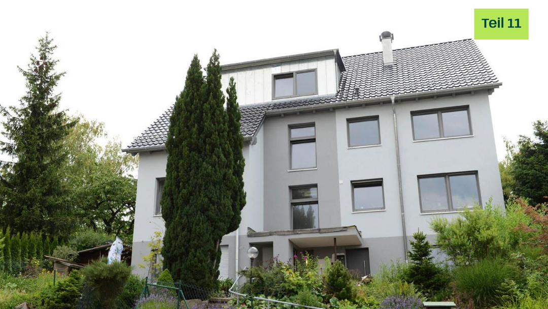 Modernes Mehrfamilienhaus mit grauer Fassade. Davor sind viele grüne Pflanzen und Büsche gepflanzt.