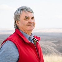 Ein Mann mit grauem Haar trägt eine rote Weste und ein blaues Hemd. Er lächelt im Freien mit einem hügeligen, trockenen Landschaftshintergrund.