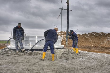 Gießen von Beton in den Fundamentkorb durch drei Arbeiter, die ein graues Rohr bewegen.