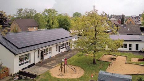 Ein zweiteiliger Gebäudekomplex mit Solaranlage auf dem Dach. Davor ist ein Spielplatz mit verschiedenen Spielgeräten und Sandplatz zu sehen.