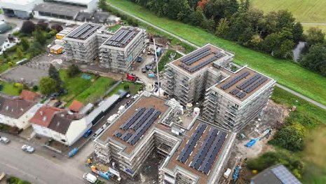 Neubauquartier mit mehreren mehrstöckigen Gebäuden im Bau. Auf allen Flachdächern sind Photovoltaikanlagen installiert. Um die Gebäude stehen Gerüste und Baumaterial, daneben liegen Wohnhäuser und Grünflächen.