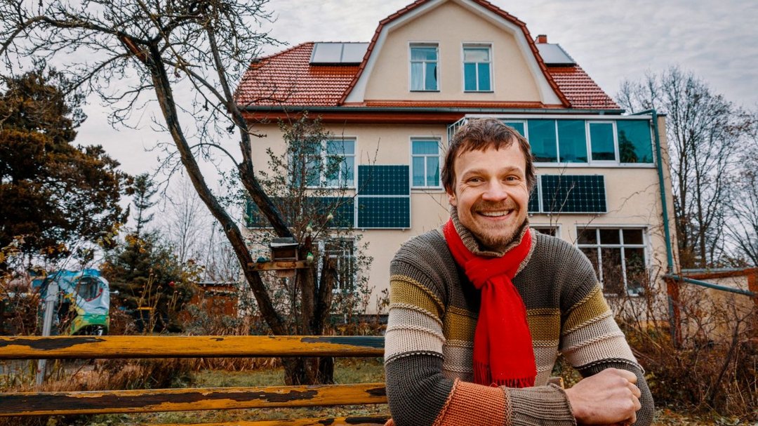 Ein Mann sitz in einem gestreiften Pullover auf einer gelben Parkbank und lächelt in die Kamera. Im Hintergrund ist ein Haus zu sehen.