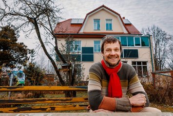 Ein Mann sitz in einem gestreiften Pullover auf einer gelben Parkbank und lächelt in die Kamera. Im Hintergrund ist ein Haus zu sehen. 