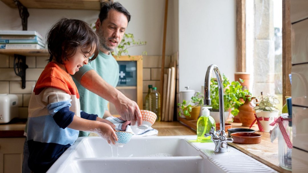 Ein Kind und ein Erwachsener spülen gemeinsam Geschirr in einer hellen Küche. Die Umgebung zeigt Pflanzen auf der Fensterbank, ein weißes Spülbecken und Seifenspender aus Plastik.